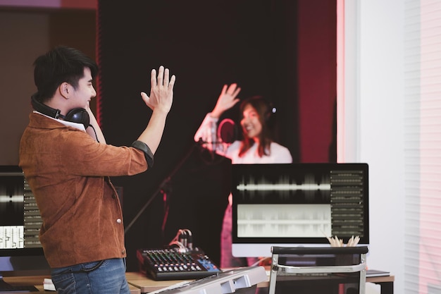 Productor asiático de pie junto a la consola de mezclas de sonido Artista compositor de música masculino feliz con antecedentes de cantante femenina