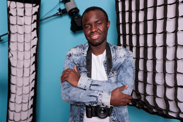 Productor de pie junto a equipos fotográficos profesionales con los brazos cruzados. Entusiasta de la fotografía posando para la sesión de fotos mientras está de pie en un estudio de producción equipado con softboxes y focos.