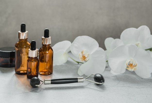 Productos para el cuidado del cuerpo y la piel, frascos cuentagotas y rodillo de masaje guasha de piedra de obsidiana en el baño con flores de orquídeas, maqueta de marca de productos cosméticos de spa