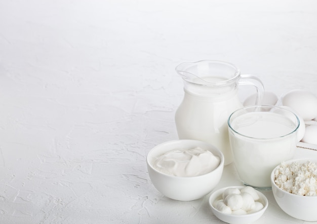 Productos lácteos frescos sobre fondo blanco de mesa. Tarro y vaso de leche, cuenco de crema agria, requesón y mozzarella. Huevos en caja de madera. Espacio para texto