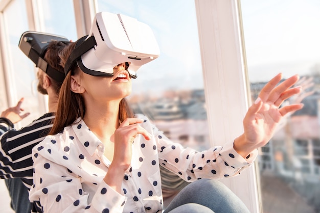 Produkt mit VR. Überraschte gut aussehende ansprechende Frau, die VR-Objekte berührt, während sie aufschaut und in der Nähe des Fensters sitzt