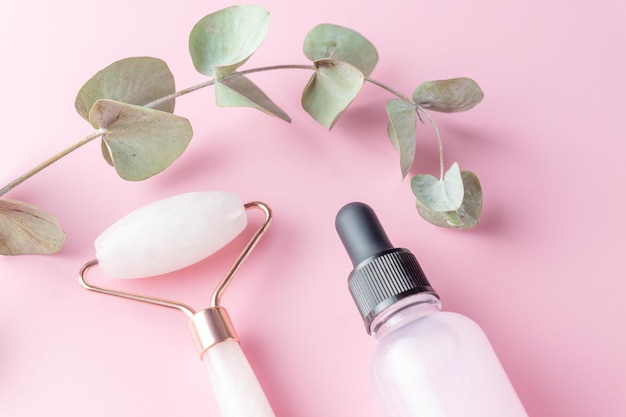 Produtos de beleza: rolo facial, óleo cosmético e soro em folhas de eqalipto com fundo rosa, vista superior. Cosméticos naturais e tratamentos de beleza.
