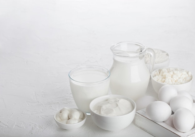 Produtos lácteos frescos no fundo da mesa branca. Jarra e copo de leite, tigela de creme de leite, queijo cottage e mussarela. Ovos em caixa de madeira. Espaço para texto