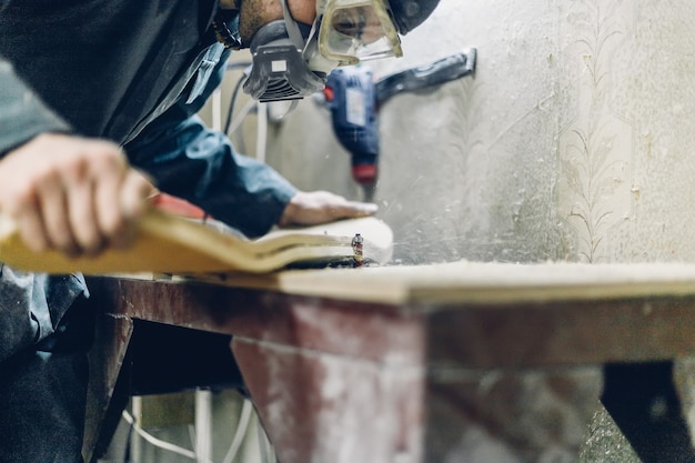 Profesión, carpintería de personas, emoción y concepto de personas - Joven artesano lijando la cubierta, reenfocando con la herramienta de lijado. fabricación de patinetas. tiene ropa protectora y máscara.