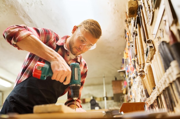 Profesión, personas, carpintería, carpintería y concepto de personas - carpintero con taladro eléctrico perforando tablones de madera en el taller