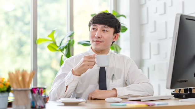 Profesional asiático exitoso hombre de negocios empleado en camisa formal con corbata sentado sostener una taza de café saludando con la mano saludar a un colega en el monitor de la computadora en el escritorio de trabajo en la oficina.