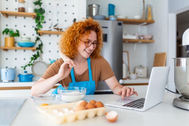 Profesional hermosa joven feliz está blogueando para su canal de cocina sobre la vida saludable en la cocina de su casa y mirando a la cámara en una computadora portátil