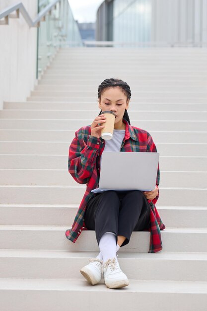 Una profesional independiente afroamericana sentada en una escalera absorta en su computadora portátil, emblemática del éxito independiente modernoxA