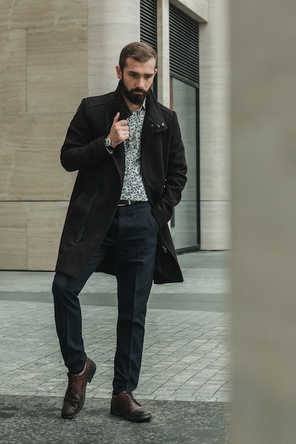 Profesional de negocios serio y reflexivo posando a toda altura frente al edificio gubernamental con su abrigo marrón y camisa ligera de moda.