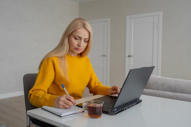 Una profesional rubia está trabajando en una computadora portátil y tomando notas en una acogedora oficina en casa