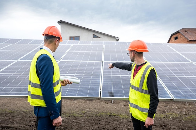 Profesionales en ropa y cascos para hombres se paran con una tableta cerca de los paneles solares recién instalados El concepto de electricidad verde en la naturaleza