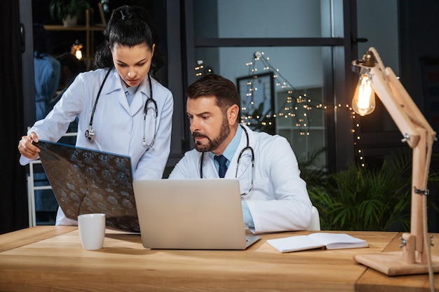 Profesionales en el trabajo. Hombre guapo con experiencia inteligente sentado frente a la computadora portátil y estudiando una foto de rayos X mientras trabaja junto con su colega