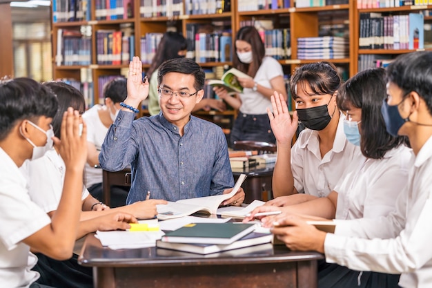 Profesor asiático levantando la mano y dando lecciones a un grupo de estudiantes universitarios para responder a la pregunta en la biblioteca y el aula de la Universidad