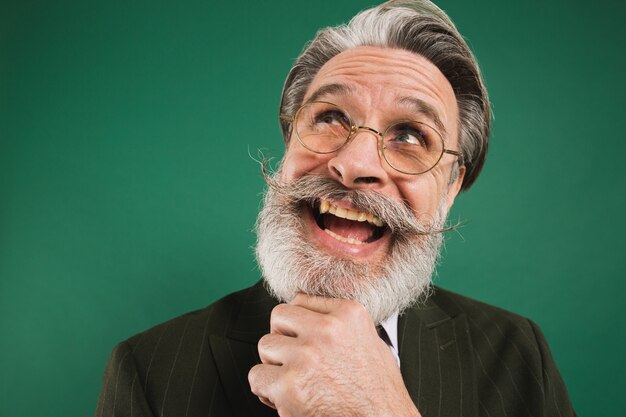 Profesor barbudo y divertido pensando y mirando hacia arriba, sosteniendo la barba en la pared verde