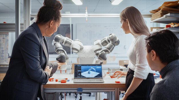 El profesor Black analiza y discute el brazo robótico futurista con dos jóvenes especialistas diversos en el laboratorio de investigación con un ingeniero portátil que ayuda a los estudiantes con el concepto del proyecto de ciencias de la computación