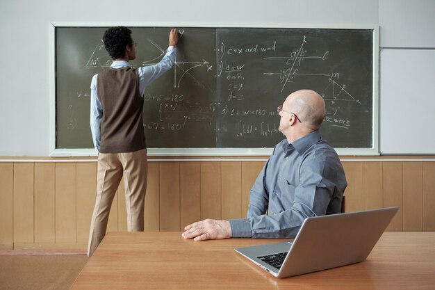 Profesor Calvo maduro sentado junto al escritorio y mirando al estudiante de pie junto a la pizarra, dibujando un gráfico y explicando sus acciones en la lección