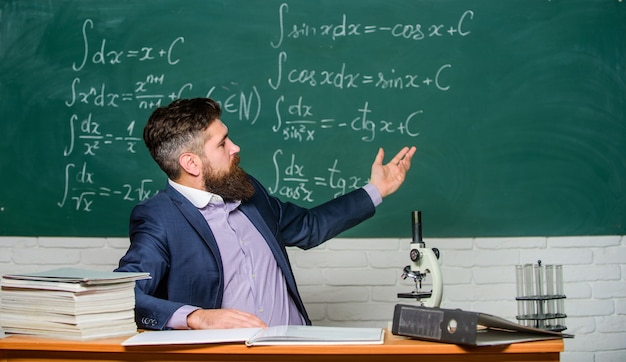 Profesor carismático hipster sentarse en el fondo de pizarra de aula de mesa. Conversación educativa. Hablar con estudiantes o alumnos. Concepto de maestro de escuela. El maestro barbudo cuenta una historia interesante.