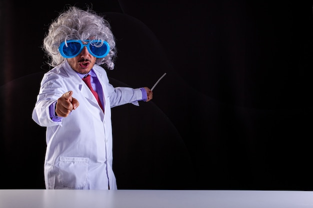 Profesor de ciencias enojado en bata blanca con el pelo descuidado en gafas divertidas puntos con el dedo sobre fondo negro