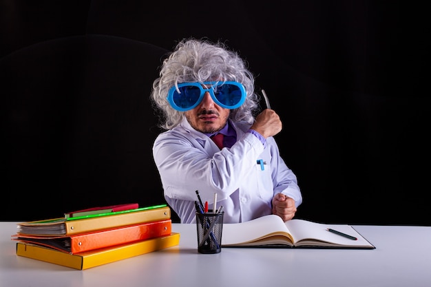 Profesor de ciencias loco en bata blanca con el pelo descuidado en gafas divertidas sentado en el escritorio sosteniendo una varita para apuntar a la pizarra