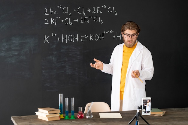 Profesor contemporáneo en bata blanca haciendo un registro de la lección de química para su audiencia en línea en la cámara del teléfono inteligente en la pizarra