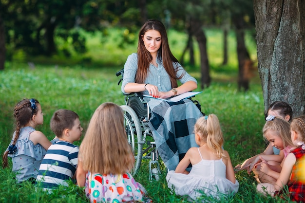 Profesor discapacitado conduce una lección con niños en la naturaleza. Interacción de un profesor en silla de ruedas con alumnos.