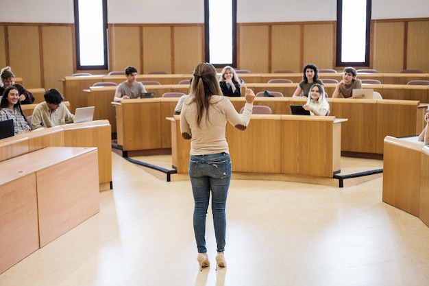 Profesor de espaldas dando clases a estudiantes universitarios en un aula con forma de anfiteatro