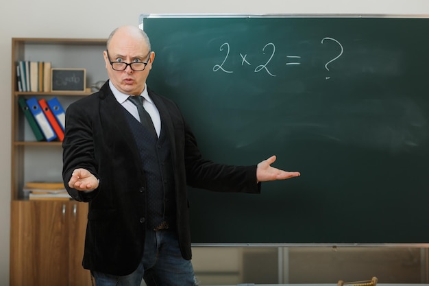 Profesor hombre con gafas de pie cerca de la pizarra en el aula explicando la lección mirando confundido extendiendo las manos a los lados