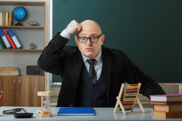 Profesor hombre con gafas con registro de clase sentado en el escritorio de la escuela frente a la pizarra en el aula mirando a la cámara sorprendido sosteniendo la mano en la cabeza