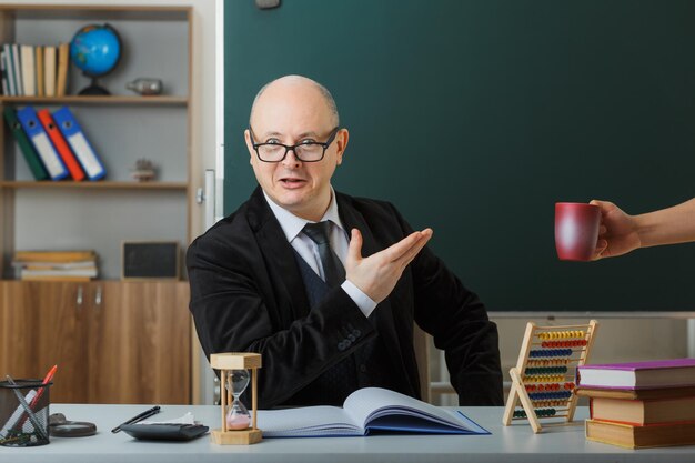 Profesor hombre con gafas sentado en el escritorio de la escuela frente a la pizarra en el aula recibiendo una taza de café con aspecto asombrado y sorprendido