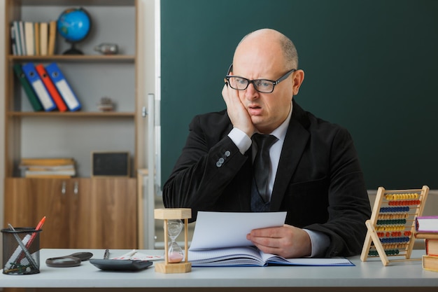 Profesor hombre con gafas sentado en el escritorio de la escuela frente a la pizarra en el aula revisando la tarea de los estudiantes que parecen preocupados