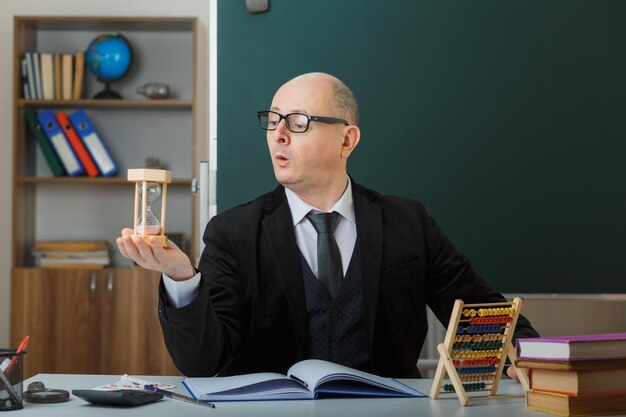 Profesor hombre con gafas sentado en el escritorio de la escuela frente a la pizarra en el aula sosteniendo un reloj de arena explicando la lección con aspecto sorprendido