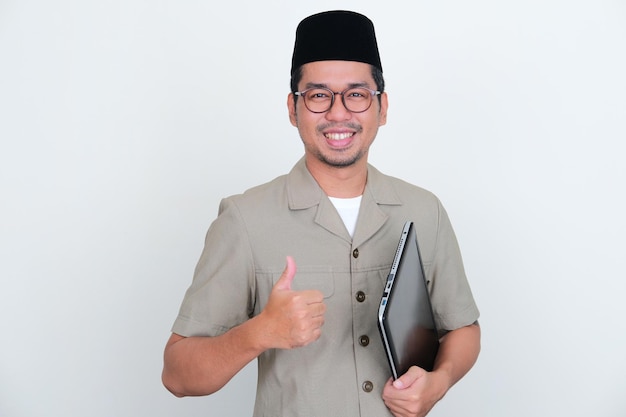 Profesor indonesio sonriendo a la cámara y levantando el pulgar mientras sostiene una computadora portátil