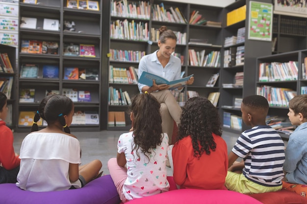 Profesor leyendo un cuento en una biblioteca para escolares sentados sobre grandes cojines de colores