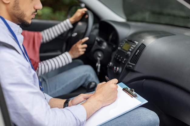 Un profesor musulmán joven y serio tomando un examen de manejo de una mujer con hiyab al volante conduciendo