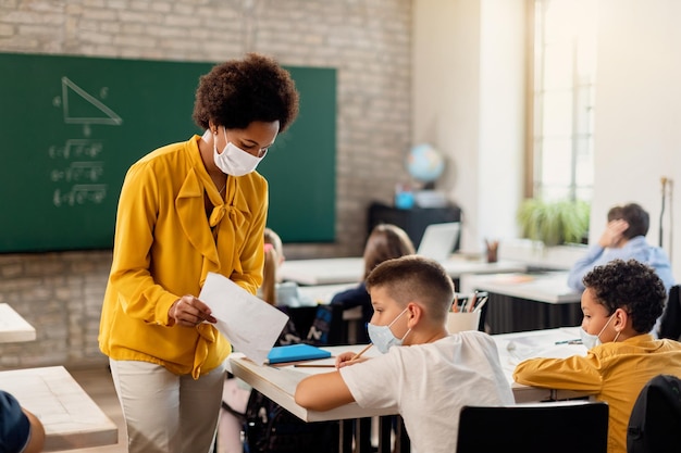 Profesor negro y estudiante de primaria con mascarillas examinando el resultado de la prueba en el aula