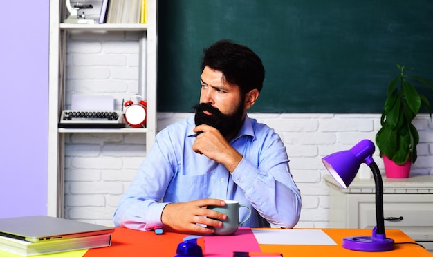 Profesor serio bebiendo café en el aula maestro de escuela sentado en el escritorio tomando un descanso para tomar café