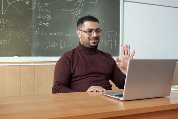 Profesor sonriente agitando la mano a su audiencia frente a la computadora portátil