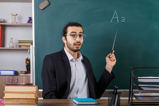 Profesor sorprendido con gafas apunta a la pizarra con un puntero sentado a la mesa con herramientas escolares en el aula