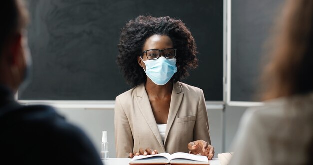 Profesora afroamericana con gafas y máscara médica sentada a la mesa en la escuela en clase, leyendo libros de texto y enseñando. Lección de literatura. Educadora haciendo preguntas a estudiantes o alumnos