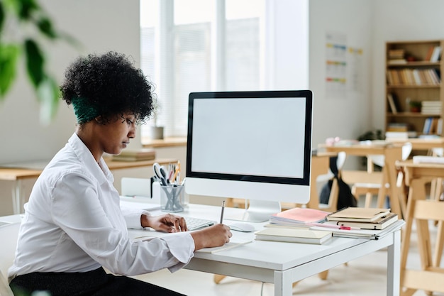 Profesora afroamericana sentada en su lugar de trabajo con computadora en clase y revisando cuadernos de s