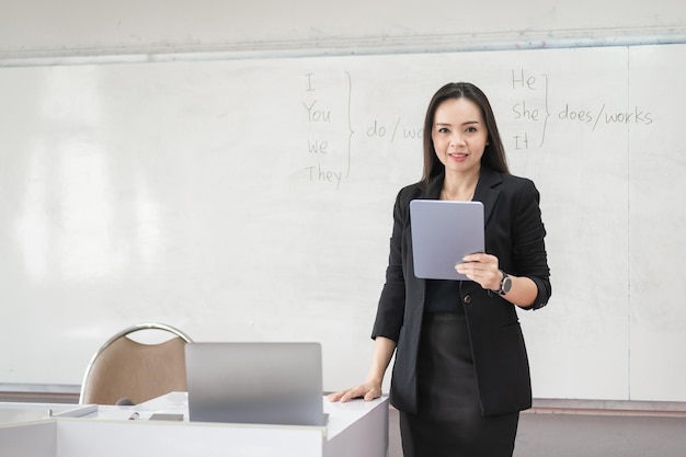 Profesora asiática alegre confiada adulta en uniforme negro del traje de negocios con la tableta digital y la enseñanza moderna en el aula de la computadora portátil