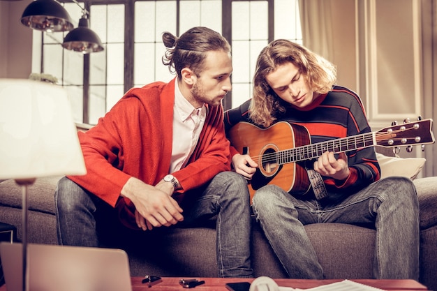 Profesora de guitarra. Profesor de guitarra de pelo oscuro y barbudo escuchando a su alumno después de practicar