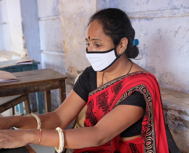 Profesora india cansada con mascarilla usando una computadora portátil en la escuela durante la pandemia de Covid-19