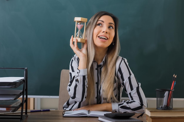 profesora joven con anteojos sentada en el escritorio de la escuela frente a la pizarra en el aula explicando la lección sosteniendo un reloj de arena mirando hacia arriba con una expresión de ensueño