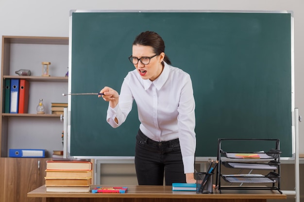 Profesora joven con gafas sosteniendo un puntero mientras explica la lección con expresión agresiva de pie en el escritorio de la escuela frente a la pizarra en el aula