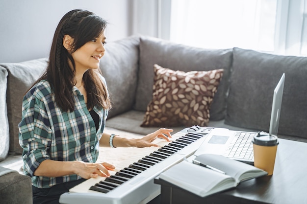 Profesora de música joven tocando el piano eléctrico enseñando de forma remota usando la computadora portátil mientras trabaja desde casa. Concepto de educación y ocio en línea.