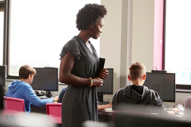 Profesora Supervisando Línea De Estudiantes De Secundaria Trabajando En Pantallas En Clase De Computación