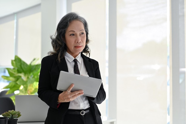 Professionelle asiatische gealterte Geschäftsfrau mit Tablet, die sich auf den Tisch stützt und in ihrem Büro arbeitet