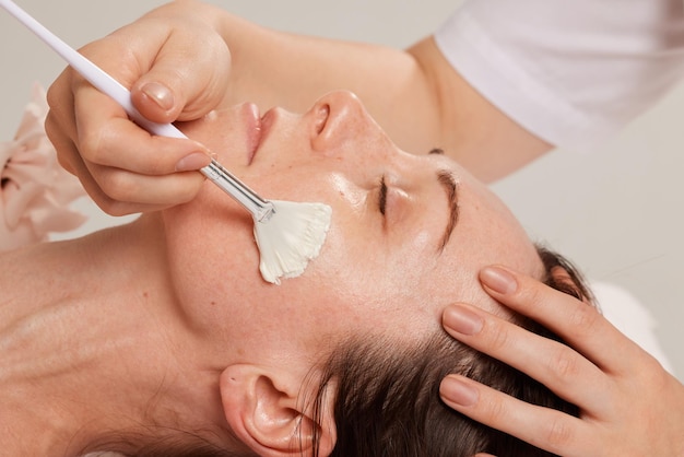 Professionelle Gesichtspflegebürste für kosmetische Eingriffe, die die Creme für das Gesicht eines Mädchens beim Termin bei einer Kosmetikerin gleichmäßig verteilt