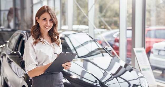 Professionelle junge Händlerin mit Zwischenablage lächelnd und kamerafreundlich betrachtend, während sie in der Nähe des modernen Automobils im Autohaus steht